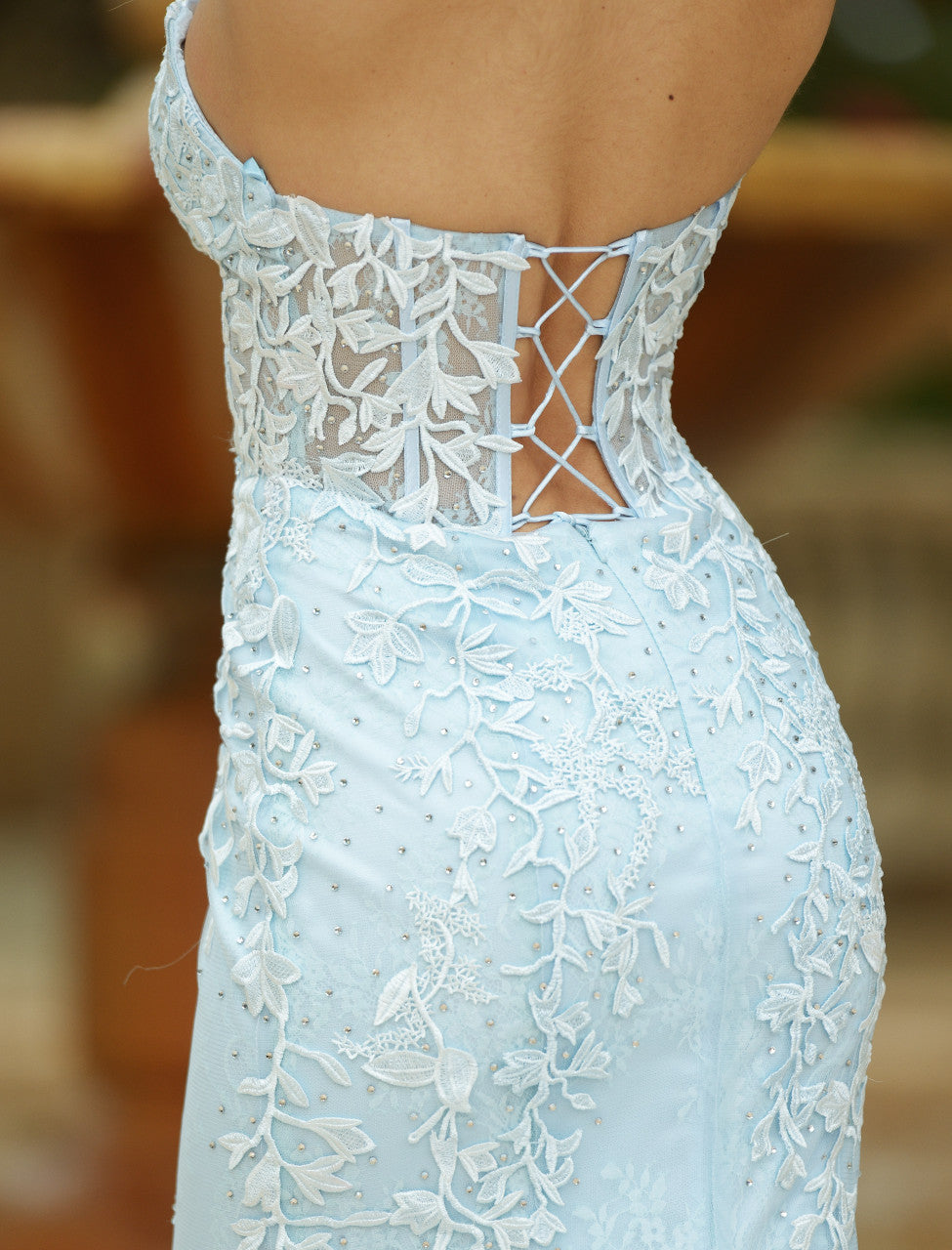 A light blue evening gown features intricate floral lace on the back and bead detailing along the fabric. The dress's lacing design adds elegance against a blurred outdoor background.