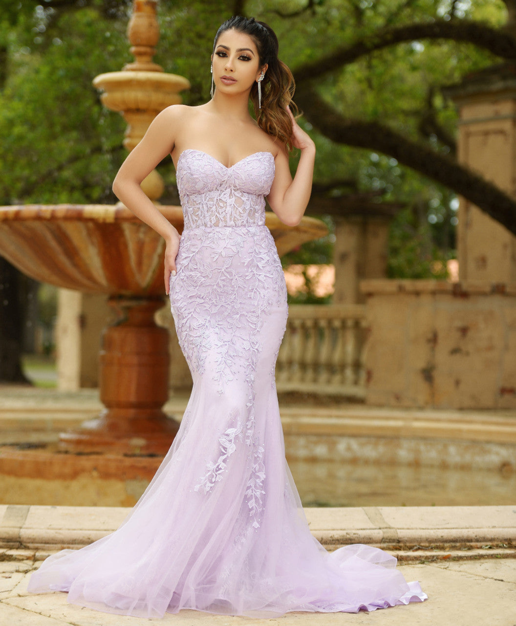 A woman stands confidently in a lavender formal gown with intricate floral designs. She poses in front of a stone fountain surrounded by greenery, exuding elegance in a serene setting.