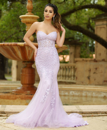 A woman stands confidently in a lavender formal gown with intricate floral designs. She poses in front of a stone fountain surrounded by greenery, exuding elegance in a serene setting.