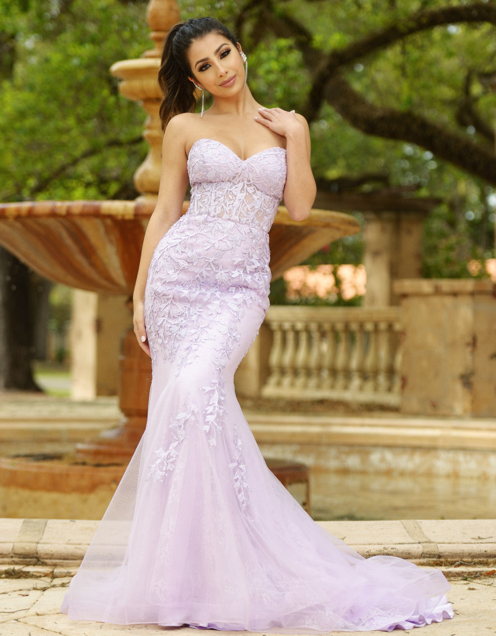 A woman in a strapless, lavender gown with floral details poses gracefully in an outdoor setting, featuring a fountain and lush greenery in the background.