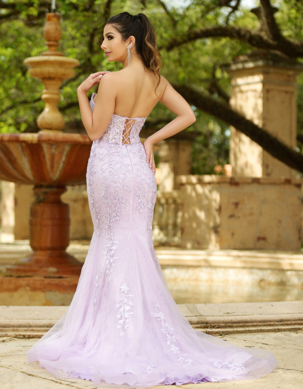 A woman in a lavender strapless gown with intricate floral embroidery poses gracefully, showcasing the gown's fitted silhouette and flowing train. She stands near a decorative fountain in a lush garden setting.