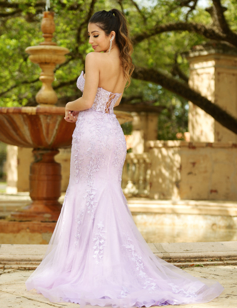 A young woman stands gracefully in a lavender gown adorned with floral details, with her back to the viewer, set against a lush garden backdrop featuring a fountain and stone structures.