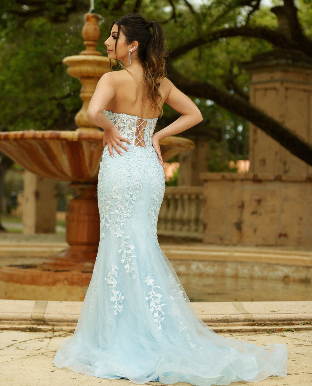 A woman in a light blue, intricately embroidered gown poses gracefully, showcasing the dress's lace details. She stands near a decorative fountain amidst a lush, garden-like environment.