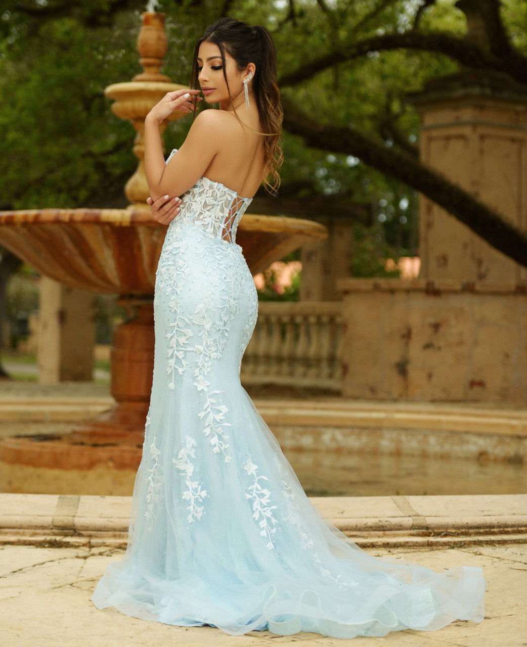 A woman in an elegant light blue gown with floral embellishments poses gracefully beside a fountain, surrounded by greenery and classical architecture, exuding beauty and sophistication.