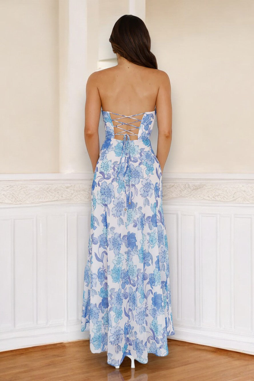 A woman in a floral blue and white strapless dress stands with her back turned. She is positioned in an interior space with light-colored walls and wooden flooring.