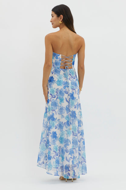 A woman is wearing a strapless, floral-printed maxi dress with blue and white hues. She is facing away, revealing a lace-up back, in a neutral background setting.