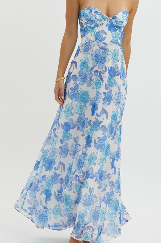 A woman wearing a strapless floral maxi dress with blue and white patterns walks gracefully. She stands against a plain background, showcasing the flowing fabric and vibrant design.