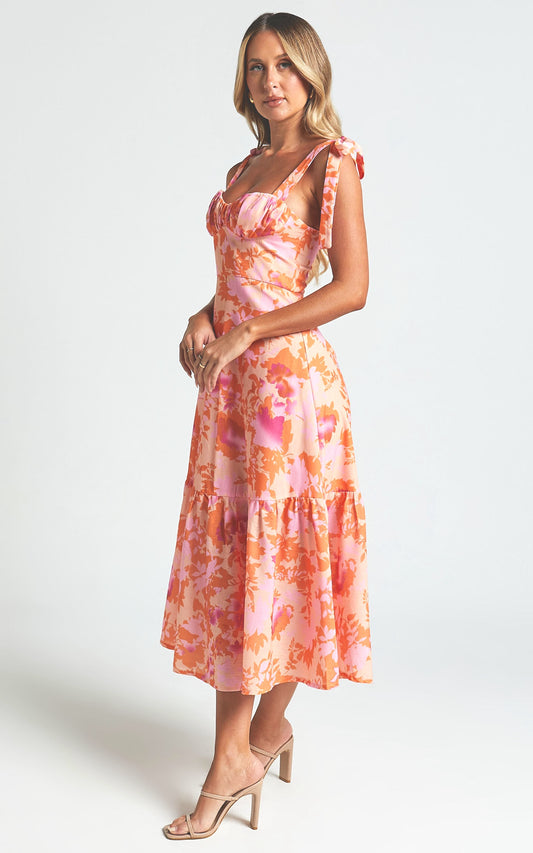 A woman stands elegantly in a floral-patterned, pastel pink and orange dress with a flared skirt and ruffled straps. She wears beige high-heeled sandals, set against a light background.