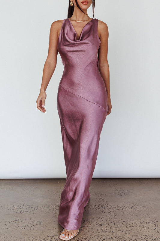 A person wears a long, flowing, mauve satin dress with a cowl neckline, walking on a textured concrete floor against a plain white background, exuding elegance and style.