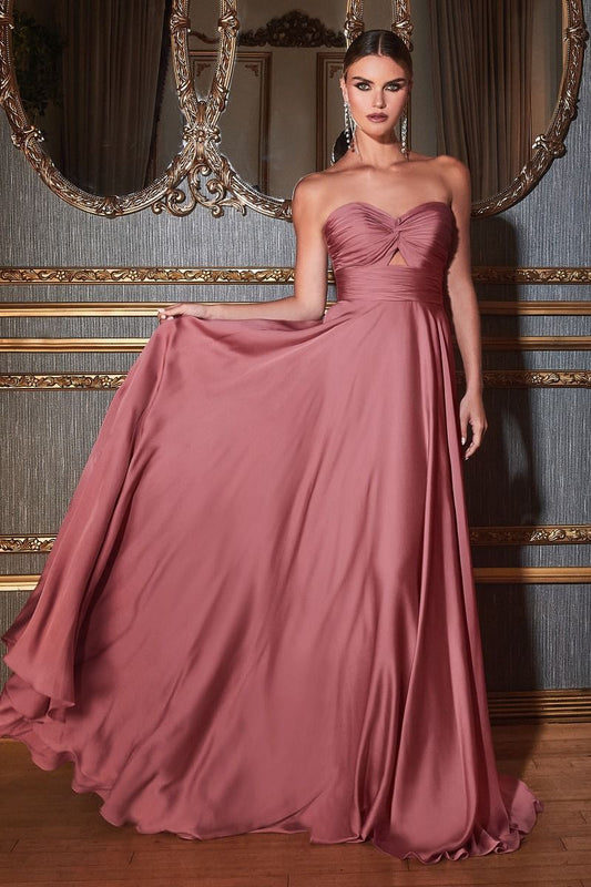 A woman wears a flowing, strapless, dusty rose gown, gracefully holding part of it as she stands against an opulent backdrop of mirrors and decorative walls.