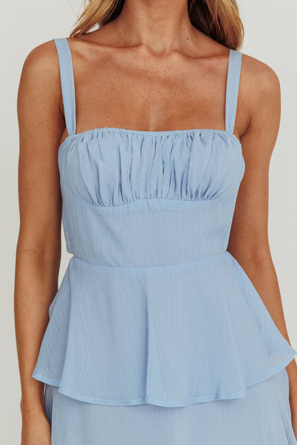 A light blue peplum top features thin straps and a gathered neckline. It is worn by a person in a neutral backdrop, emphasizing the soft texture and color of the fabric.