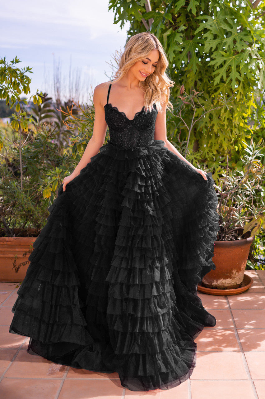 A woman in a ruffled black gown smiles while holding the dress. She's standing on a tiled surface surrounded by greenery and potted plants, with a bright sky in the background.