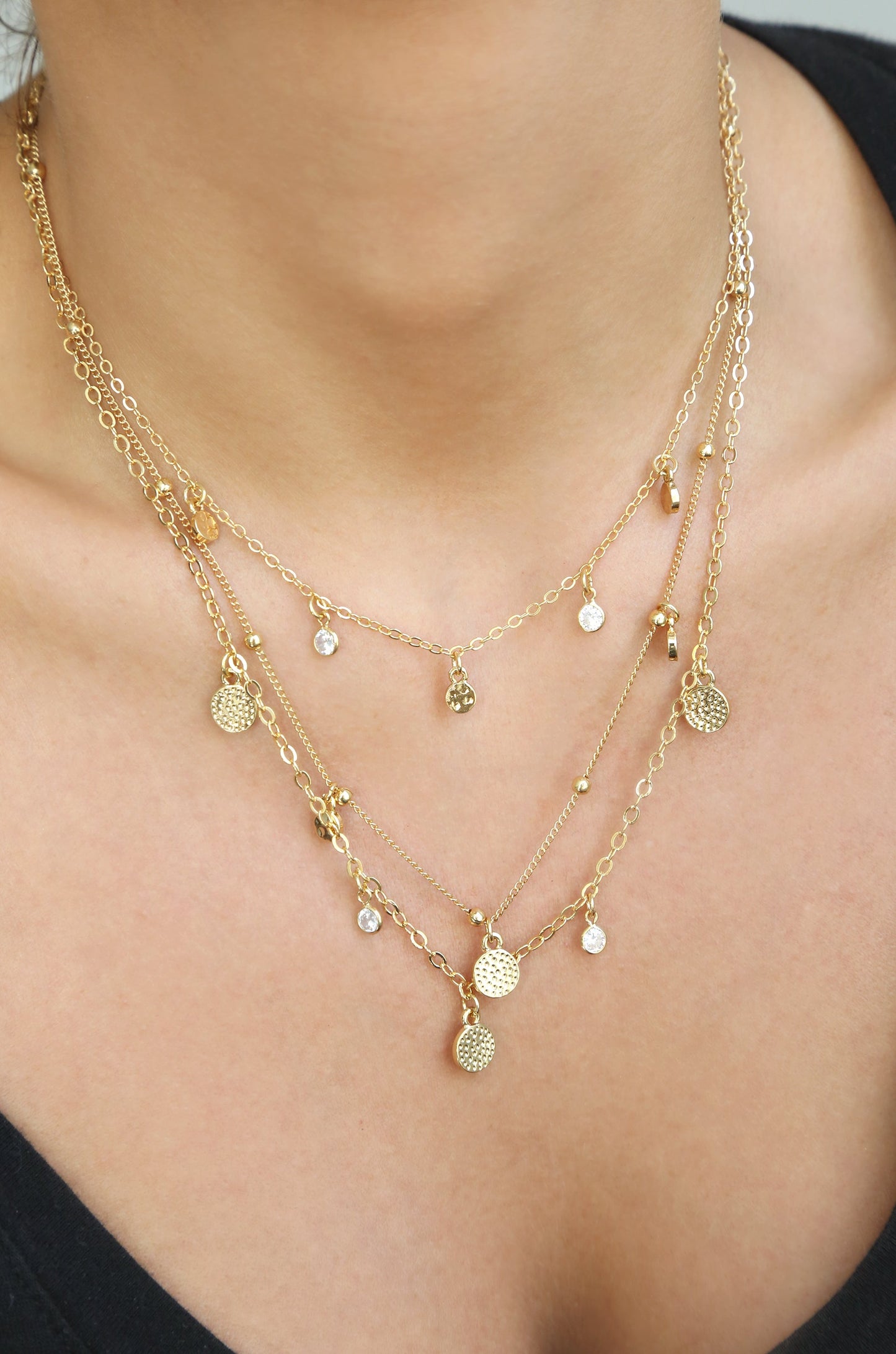 A layered gold necklace adorned with small charms and clear crystals rests on a personâ€™s neck, enhancing the collarbone above a dark top in a softly lit setting.