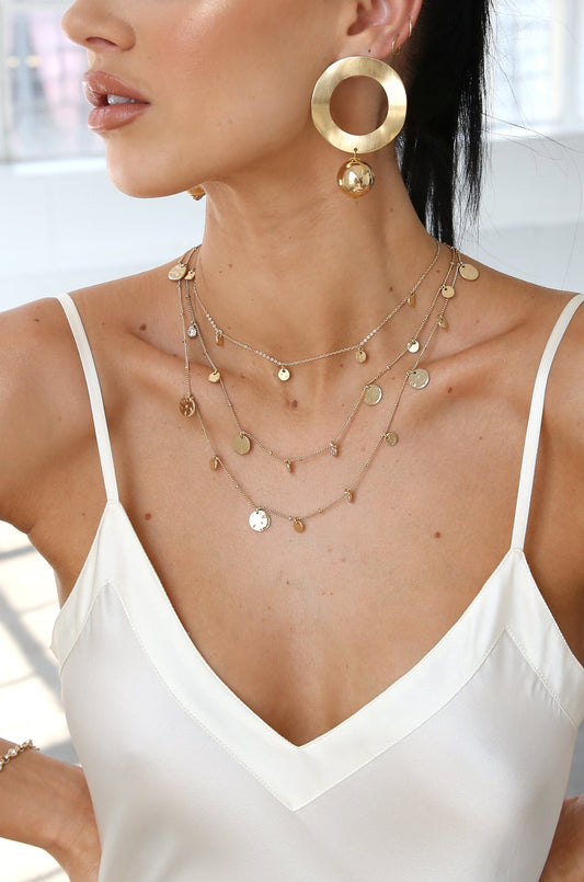 A woman wears layered gold necklaces and large hoop earrings, showcasing her accessories against a bright, neutral background. The focus is on her jewelry and elegant neckline.