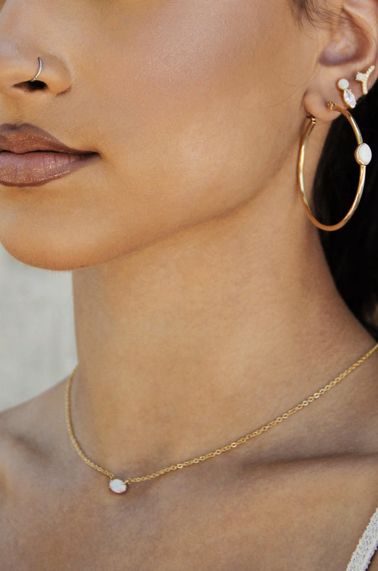 A close-up of a womanâ€™s profile showcases her gold hoop earrings and a delicate gold necklace with an opal pendant, set against a softly blurred background.