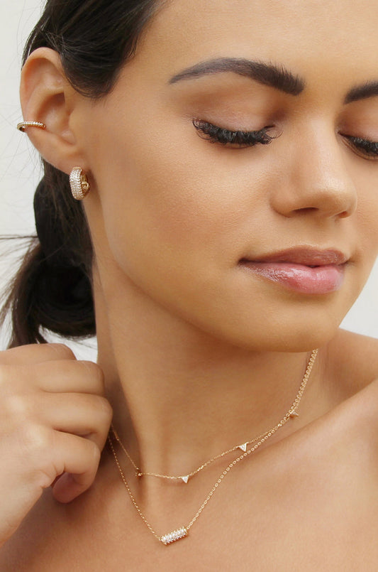 A young woman showcases various gold jewelry pieces, including earrings and layered necklaces, as she lightly touches her neck, set against a neutral background. Her expression is serene and contemplative.