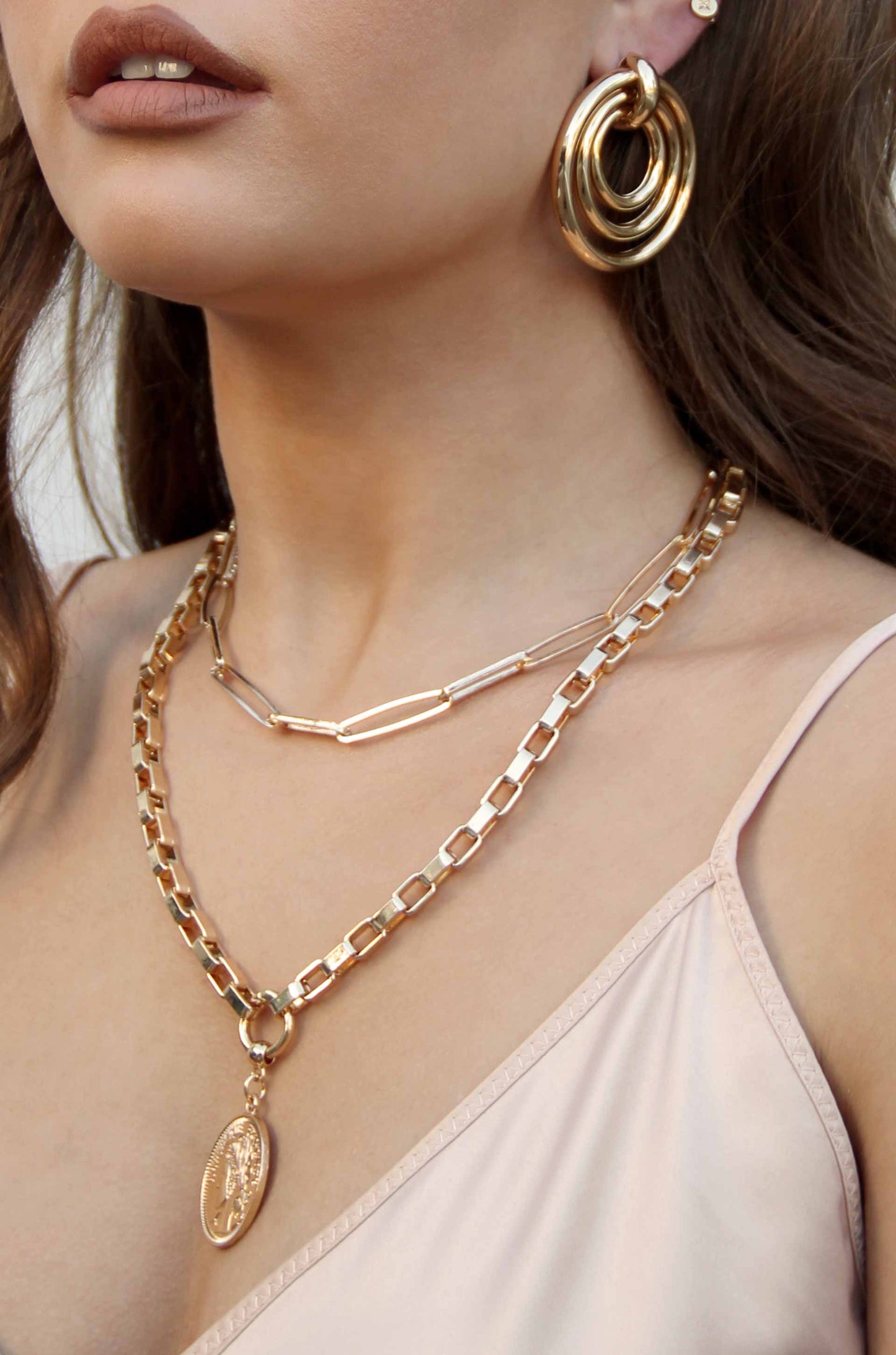 A close-up of a person's neck adorned with layered gold necklaces and large hoop earrings. The background is neutral, highlighting the jewelry and the person's skin tone.