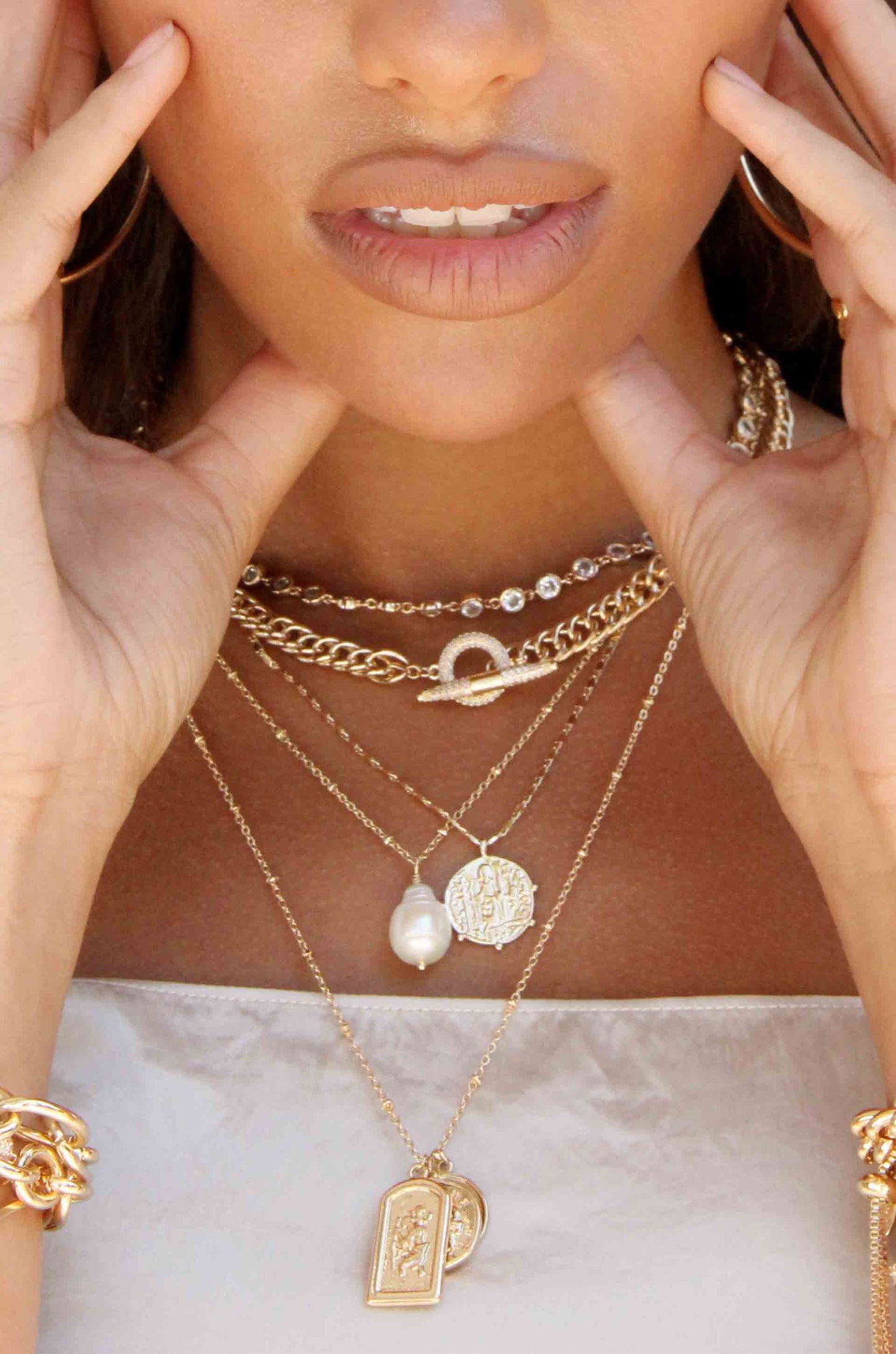 A woman gently touches her cheeks with both hands while wearing multiple layers of gold necklaces, including chains and pendants, against a soft, light-colored fabric backdrop.