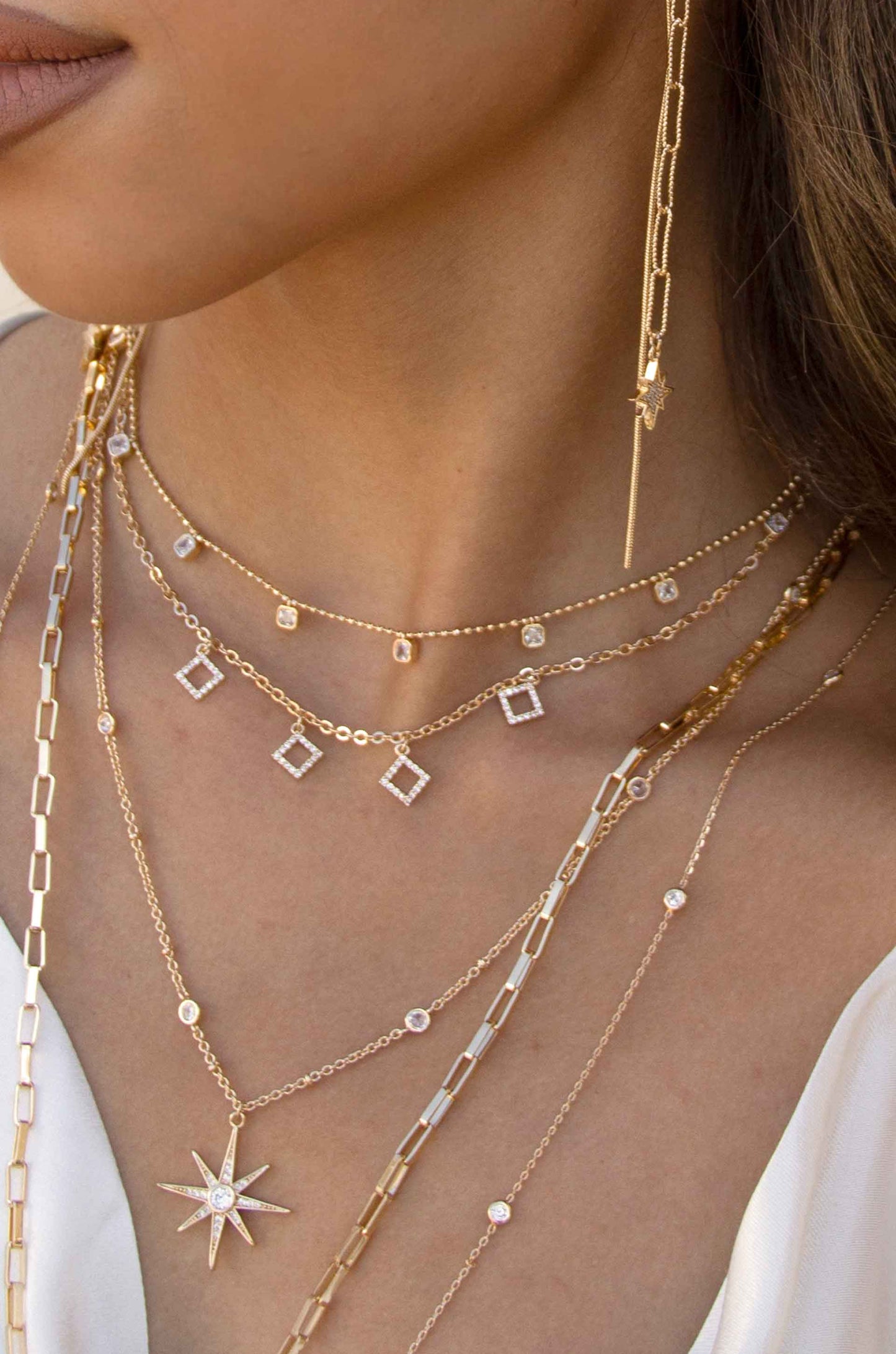 Multiple strands of delicate gold necklaces drape across a person's neckline, featuring various shapes and sparkling elements. One long earring extends downward, adding elegance to the look.