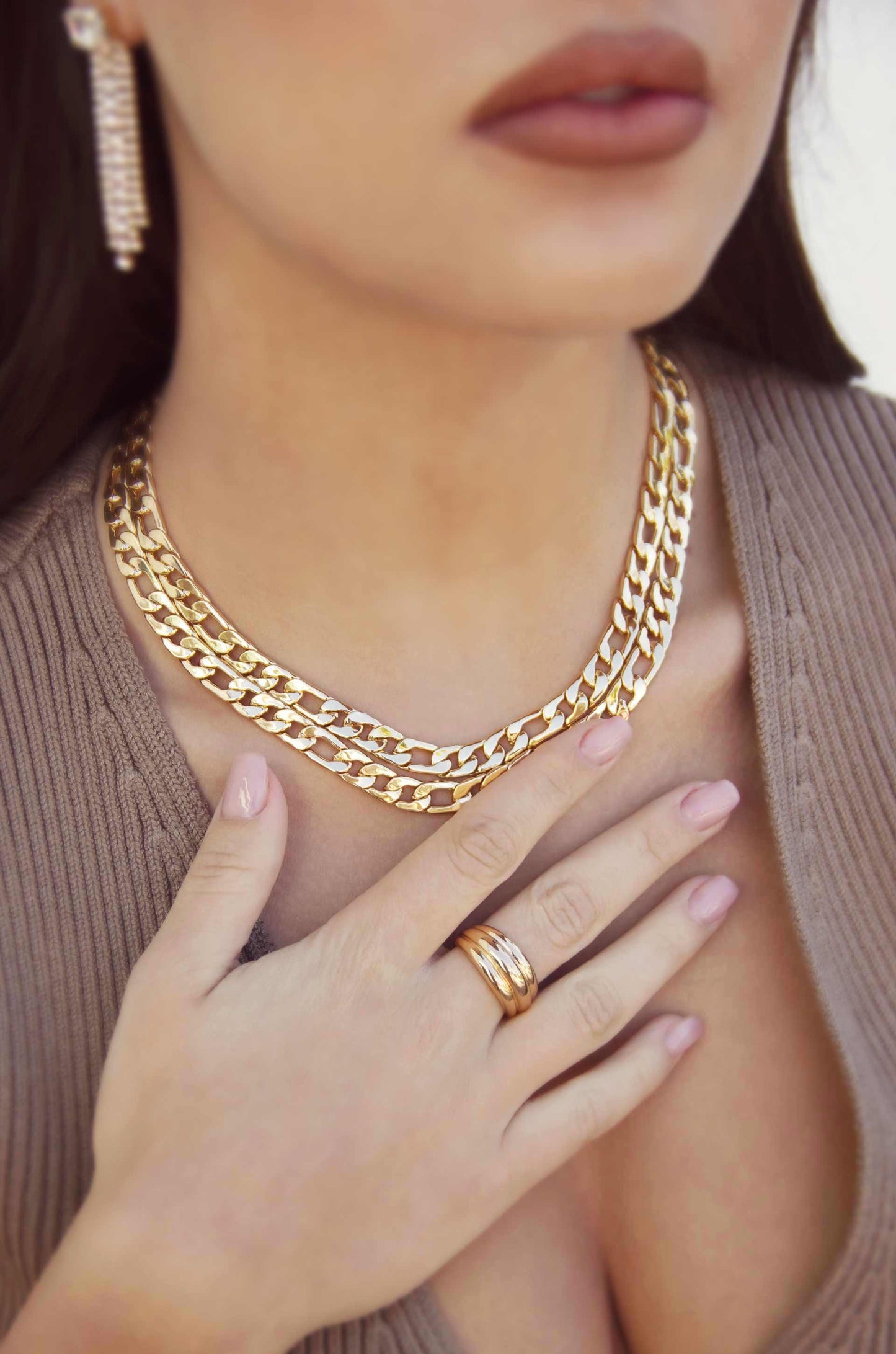 A close-up of a woman wearing multiple gold chains around her neck, gently touching one with her hand, while showcasing a gold ring on her finger, against a neutral background.