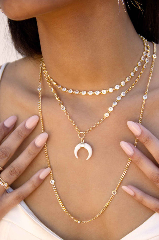 A person wears layered gold necklaces featuring circular and crescent-shaped pendants. They touch the chains with one hand against a neutral background, showcasing their polished, light pink nails.