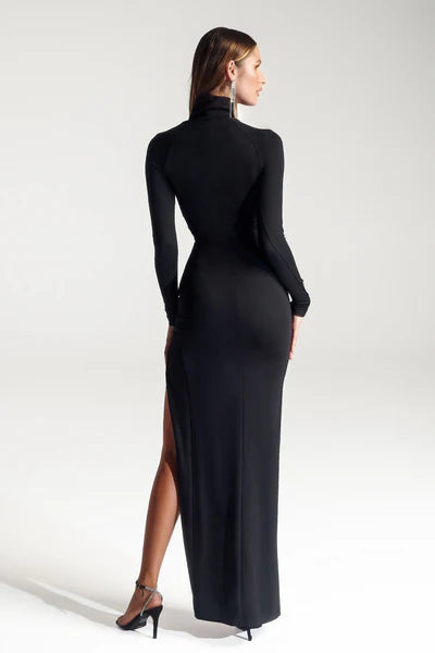 A woman wears a form-fitting black dress with a high neckline and long sleeves, showcasing a side slit. She stands against a neutral background, emphasizing elegance and style.