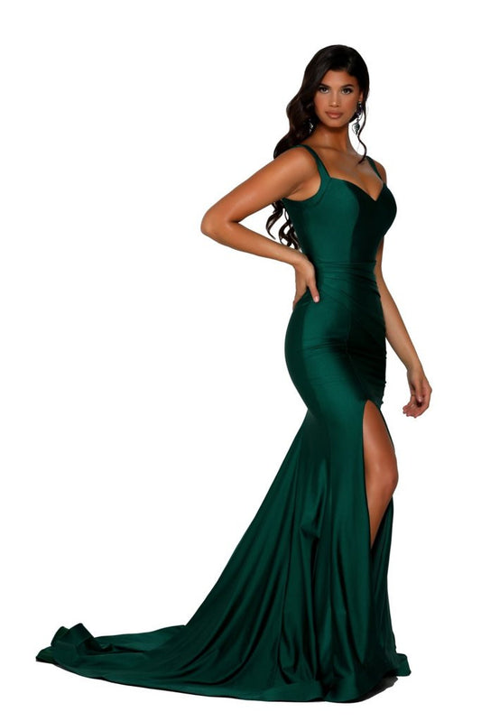 A woman wearing a form-fitting dark green evening gown poses confidently, showcasing a thigh-high slit. The dress flows dramatically, creating an elegant silhouette against a plain background.