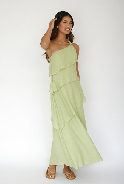 A woman stands smiling in a flowing, layered, light green dress with one shoulder strap. She has long, dark hair and is positioned against a plain white background, exuding a relaxed, happy vibe.