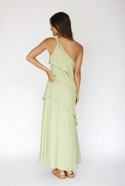 A woman stands with her back to the viewer, wearing a flowing, one-shoulder, light green maxi dress that features layered ruffles. She has long, straight hair and is positioned against a plain white background.