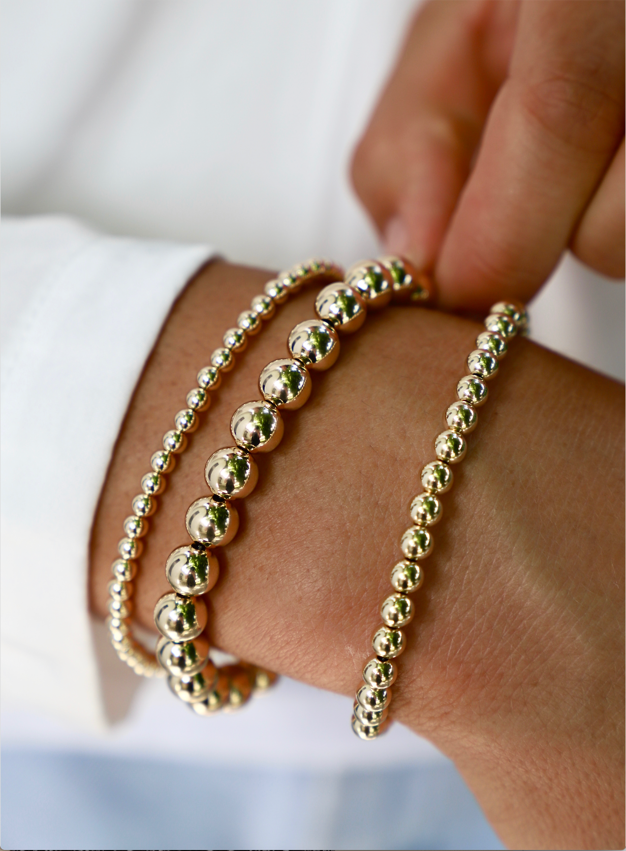 Golden bead bracelets adorn a hand, with the fingers adjusting the placement. The background features a blurred white shirt, suggesting a casual yet elegant setting.