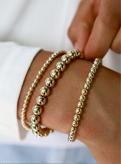 Golden bead bracelets adorn a hand, with the fingers adjusting the placement. The background features a blurred white shirt, suggesting a casual yet elegant setting.