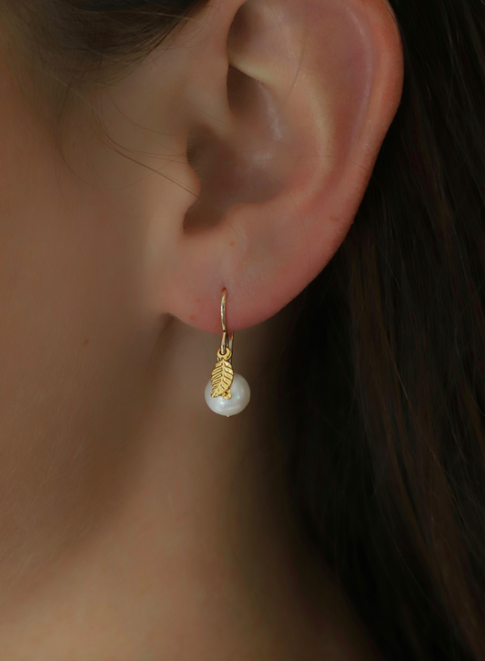 A gold earring featuring a leaf design and a pearl hangs from the lobe of an ear, set against a blurred background, highlighting the accessory's elegance.