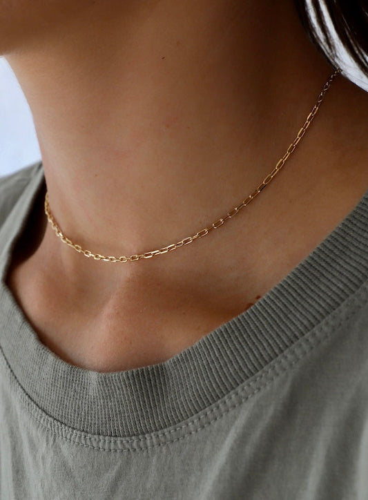 A delicate gold chain necklace adorns a person's neck, reflecting light subtly. The wearer is dressed in a simple, slightly wrinkled olive green shirt, emphasizing a casual, understated style.