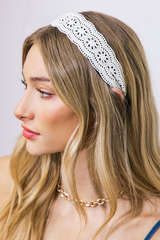 A woman with long, wavy blonde hair wears a decorative white headband and a gold chain necklace, showcasing a side profile against a soft, neutral background.