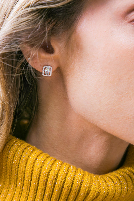 A close-up of a woman's ear adorned with a sparkling rectangular earring. The woman is wearing a vibrant yellow sweater with ribbed texture, set against a softly blurred background.