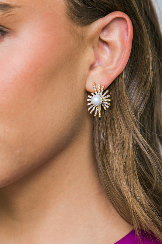 A close-up of a woman's ear adorned with a starburst-shaped earring featuring a central pearl and sparkling accents, set against her smooth, lightly tanned skin and shoulder-length hair.