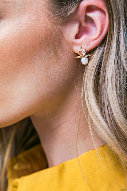 A woman's ear is adorned with a decorative earring featuring a pearl and faceted stone, complemented by a golden design. Her blonde hair softly frames her neck, and she wears a yellow garment.