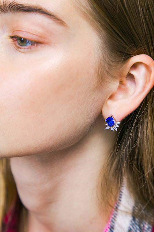 A close-up of a woman's ear features a large, rectangular blue gemstone earring adorned with smaller, sparkling elements. The background is softly blurred, emphasizing the jewelry and the woman's natural complexion.