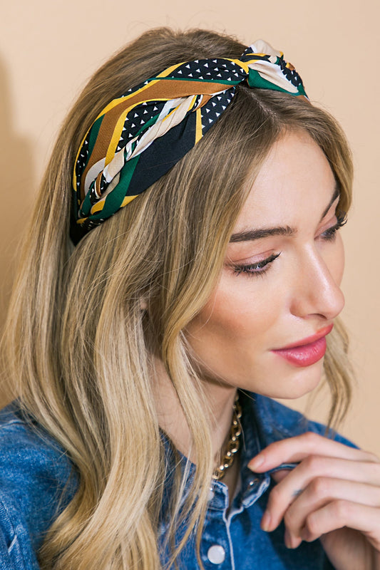 A woman with long, wavy blonde hair wears a patterned headband and a denim shirt. She is looking thoughtfully to the side against a soft, neutral background.