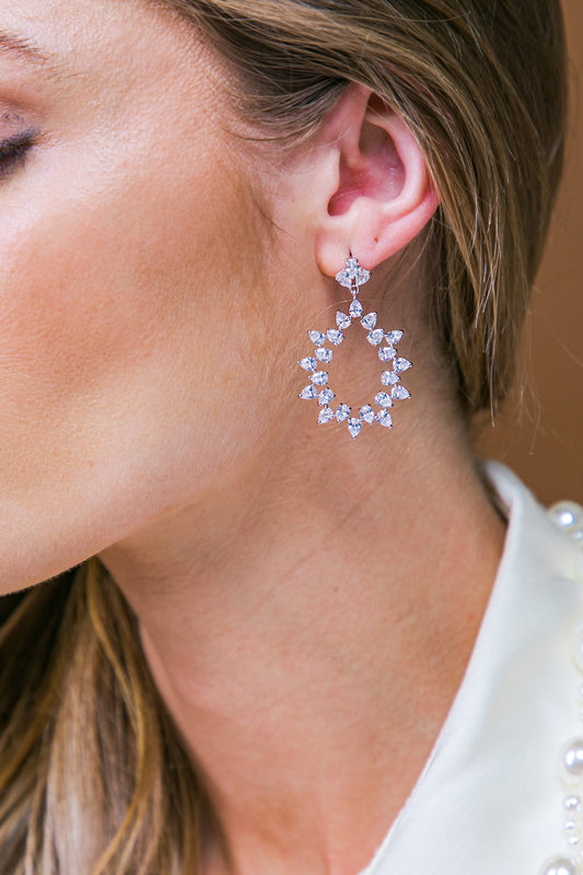 A sparkling, intricate earring featuring pear-shaped and round stones hangs elegantly from a woman's ear, showcasing her smooth skin and flowing hair against a neutral-colored background.