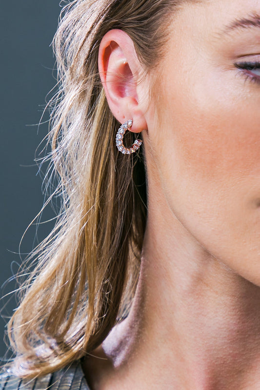 A close-up of a woman's ear showcases a sparkling silver hoop earring adorned with small crystals, with her wavy, light brown hair cascading nearby, and a neutral background.