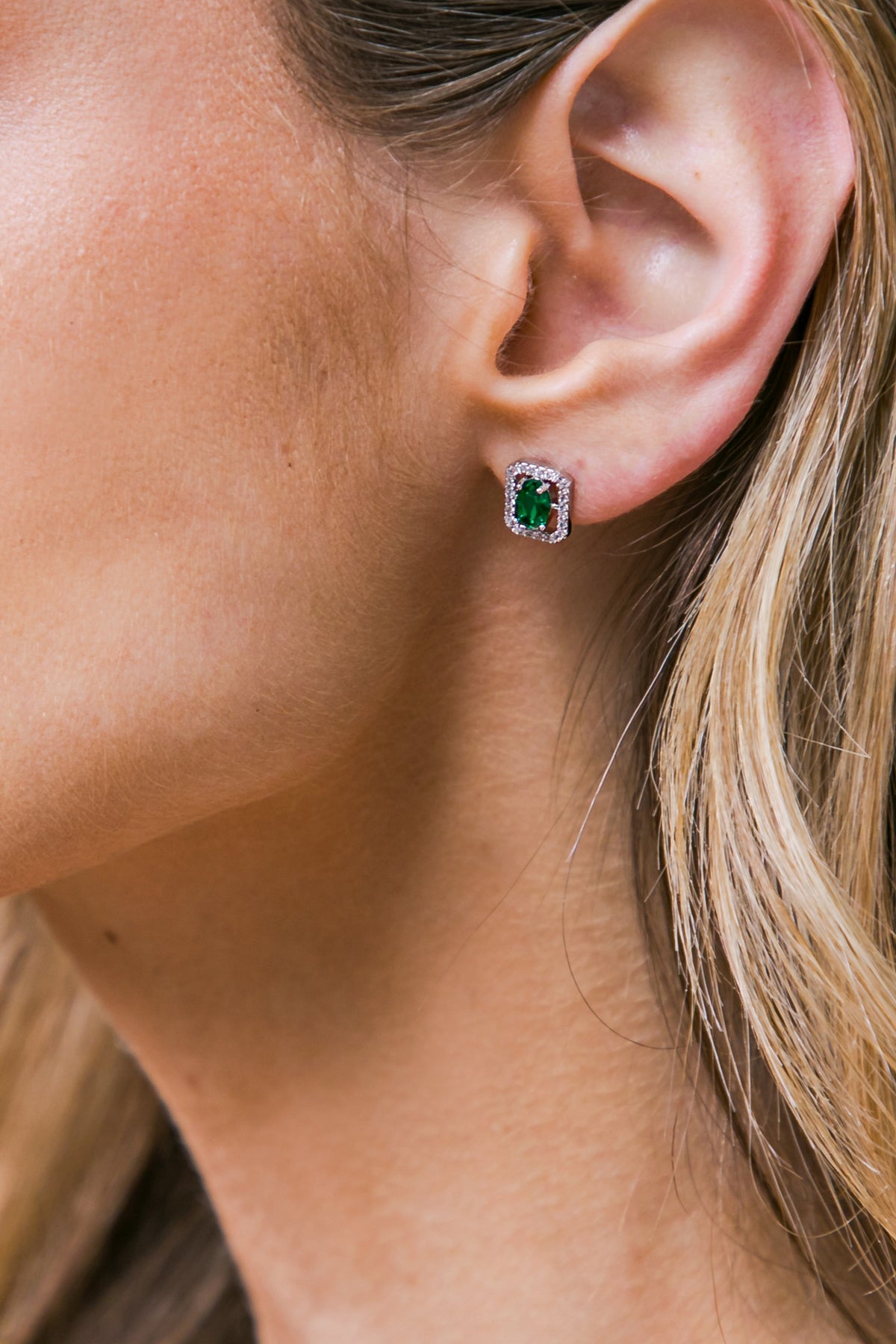 A close-up of an ear featuring an elegant earring. The stud earring has a green gemstone surrounded by a sparkling border, set against a backdrop of smooth skin and flowing hair.