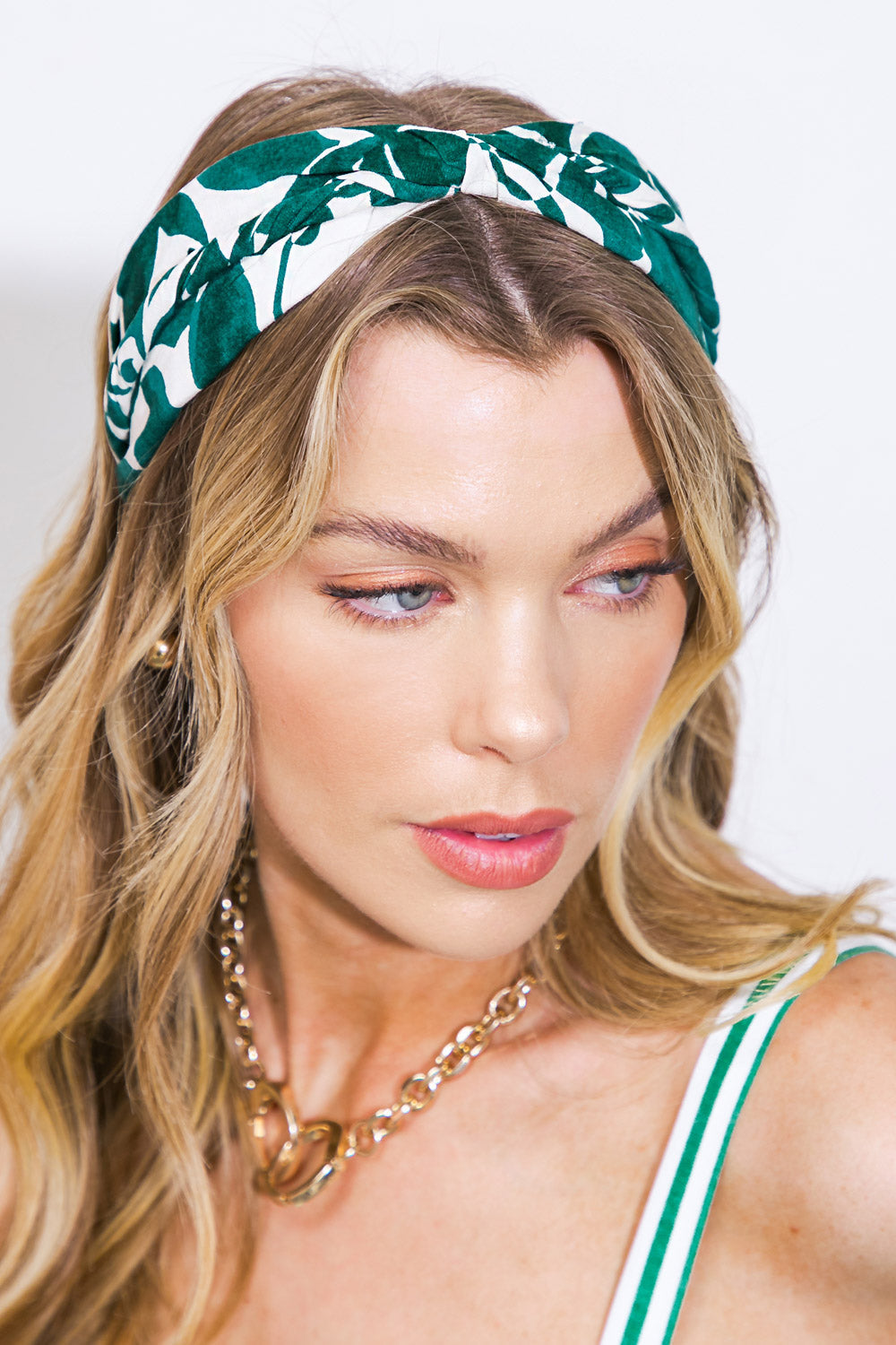 A woman with long blonde hair wears a floral green and white headband and a gold chain necklace, gazing slightly downwards against a plain, bright background.