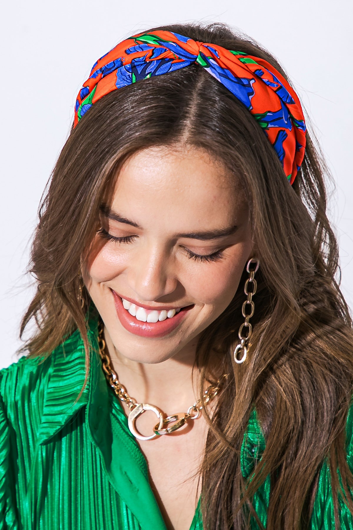 A woman with wavy brown hair wears a colorful floral headband and a green pleated top, smiling and looking downward in bright, well-lit surroundings. Gold chain earrings add to her outfit.