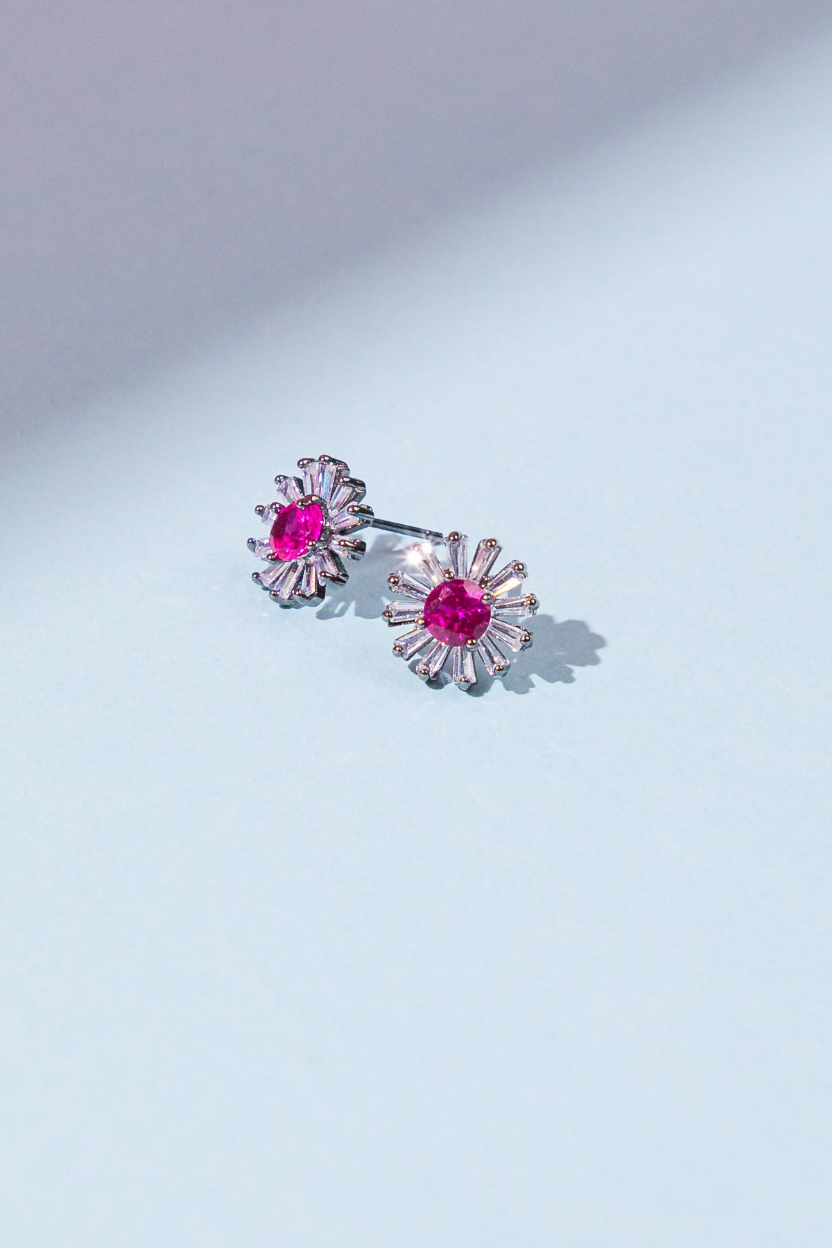 Two decorative earrings with vibrant pink gemstones and silver accents rest on a light blue background, reflecting soft light and casting subtle shadows.