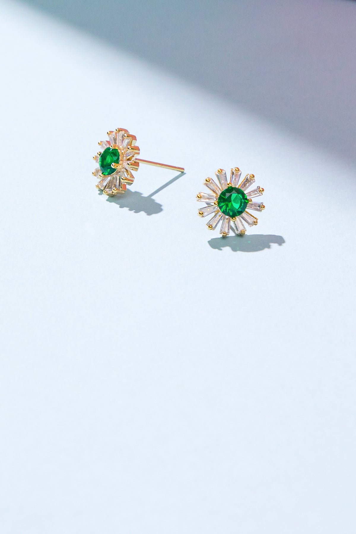 Two gold-tone earrings with green oval center stones and radiating clear accents are displayed on a light blue surface, illuminated by soft light, casting subtle shadows.