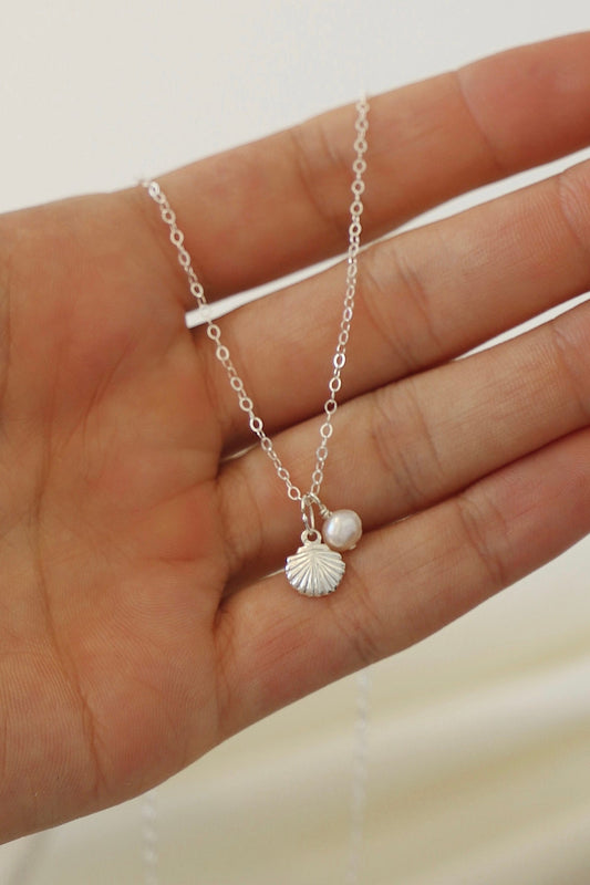 A hand holds a delicate silver necklace featuring a shell charm and a small pearl, showcasing the jewelry against a softly blurred light background.