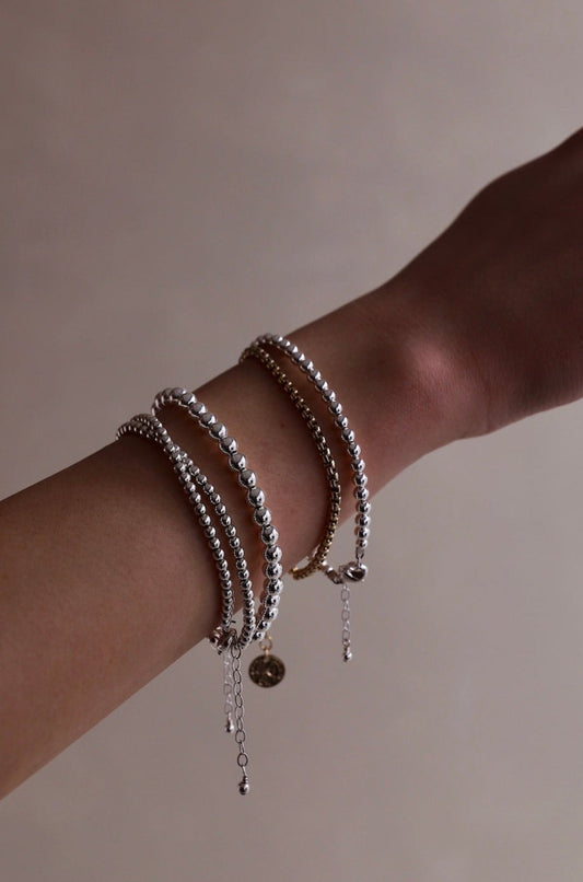 A hand with several bracelets rests against a neutral background. The bracelets are silver and gold with varying beads and chains, adding depth and texture to the wrist.