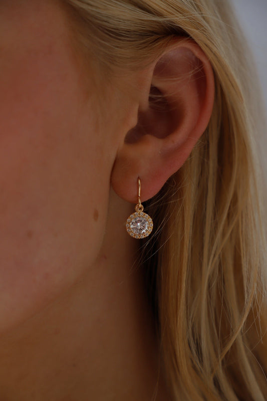 A gold hoop earring hangs from a person's ear, showcasing a large sparkling gem in the center. The person's blonde hair softly frames the ear against a blurred, light background.