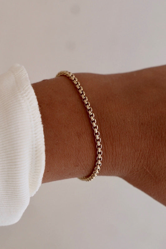 A gold chain bracelet is worn on a wrist. The arm is partially visible, with the fabric of a white long-sleeve shirt cuff peeking out, set against a light background.